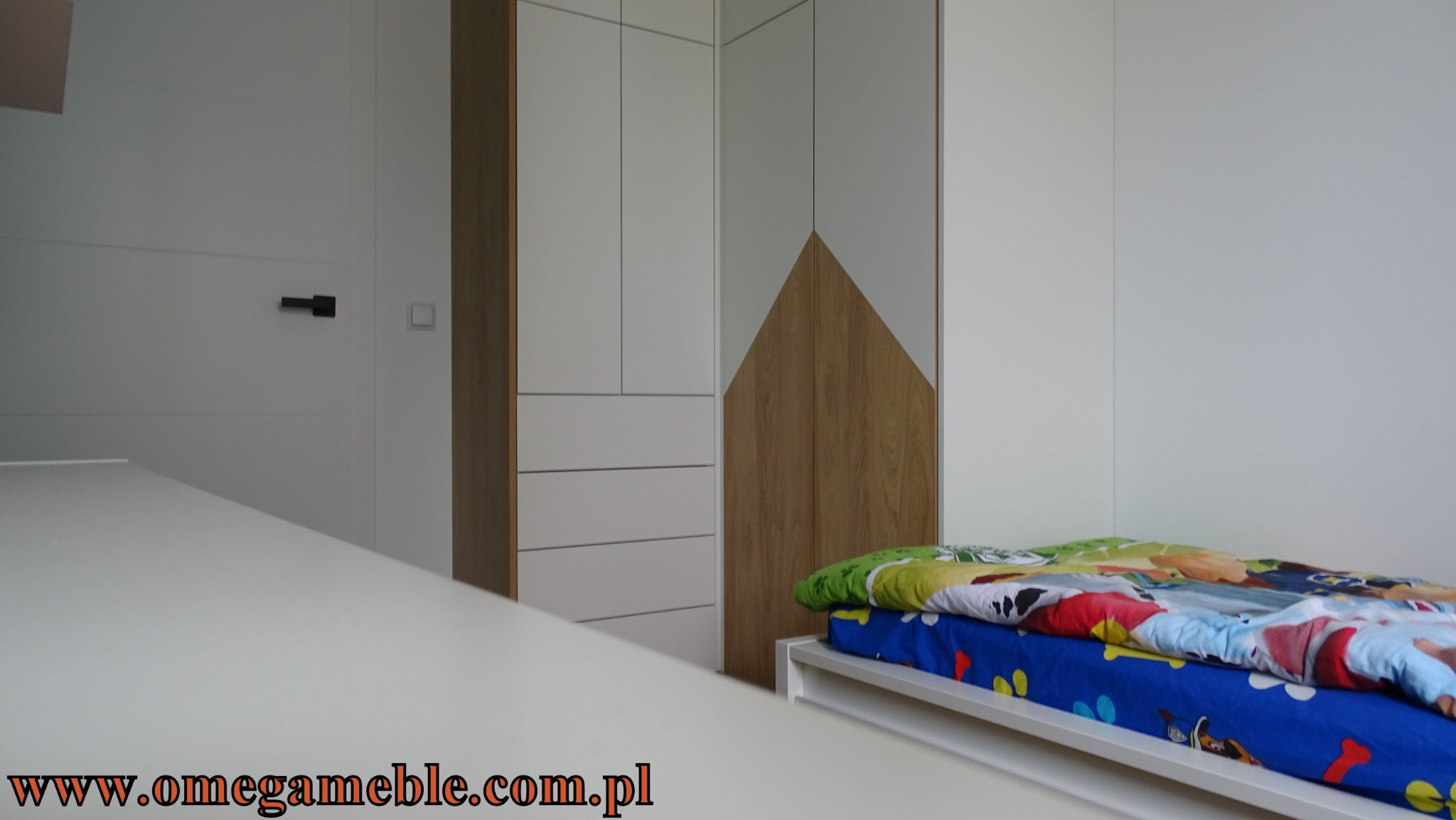 Biała szafa z drewnianymi akcentami w pokoju dziecięcym, widoczna część łóżka z pościelą w bajkowe wzory, minimalistyczny design.