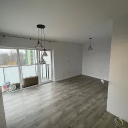 Pusty pokój w Bezrzeczu z panelami podłogowymi ułożonymi w jodełkę, trzema wiszącymi lampami w stylu industrialnym i balkonem z widokiem na drzewa.