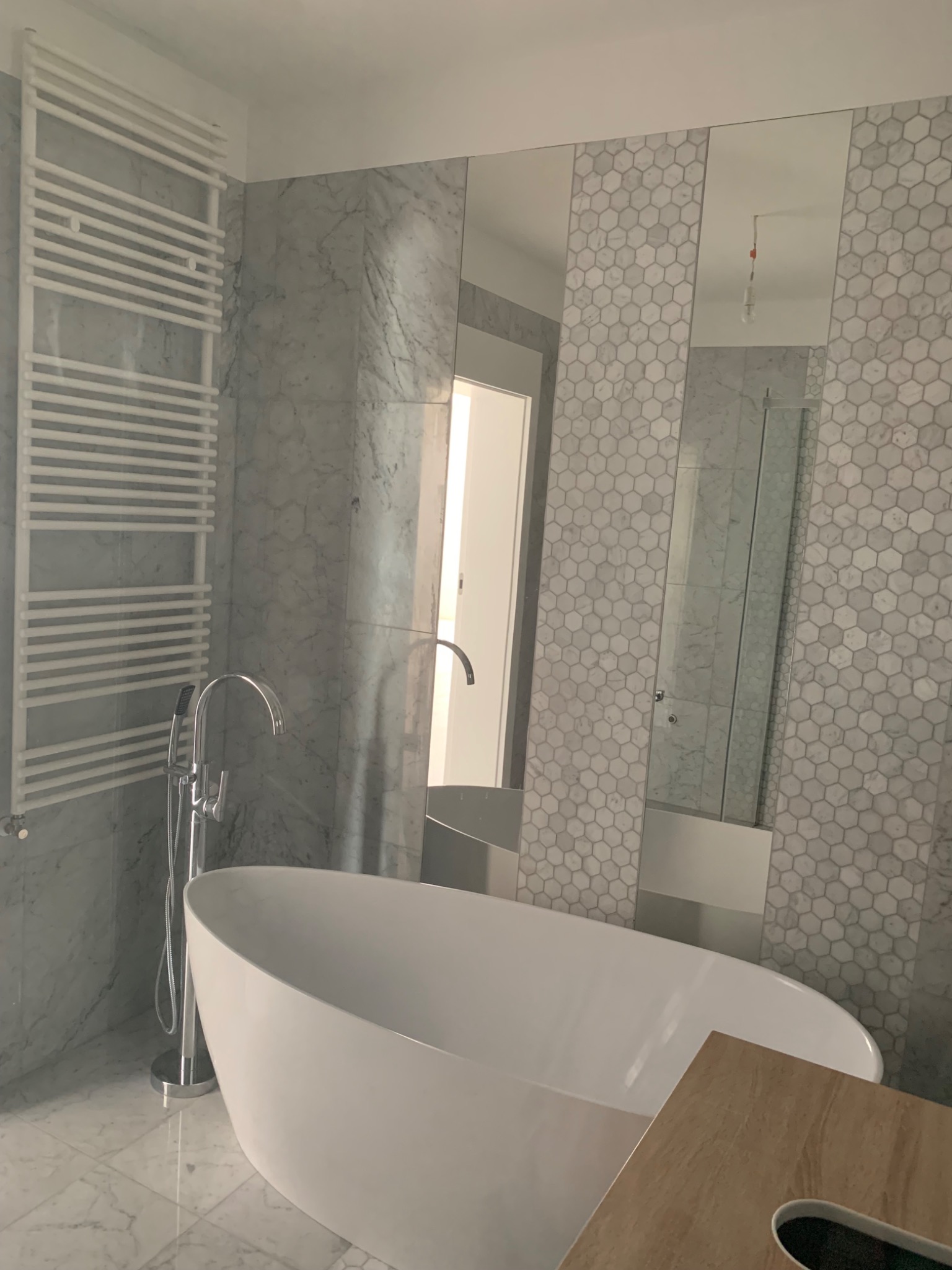 Nowoczesna łazienka z wolnostojącą wanną, chromowaną armaturą, grzejnikiem drabinkowym i ścianami wyłożonymi marmurowymi płytami oraz mozaiką heksagonalną, widok na lustro i kabinę prysznicową w tle.