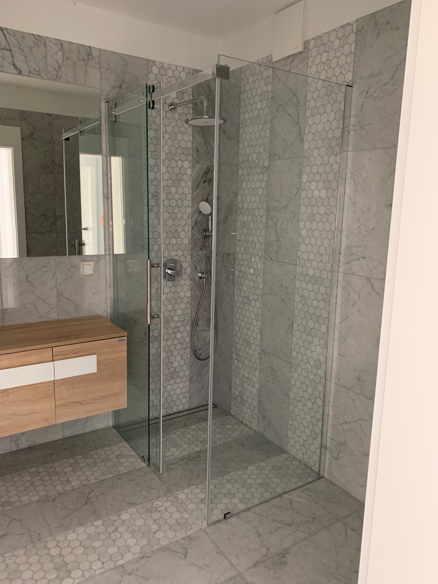 Nowoczesna kabina prysznicowa ze szklanymi ścianami, wykończona marmurowymi płytkami w odcieniach szarości i bieli, z elementami mozaiki heksagonalnej. Drewniana szafka pod umywalką z białymi...