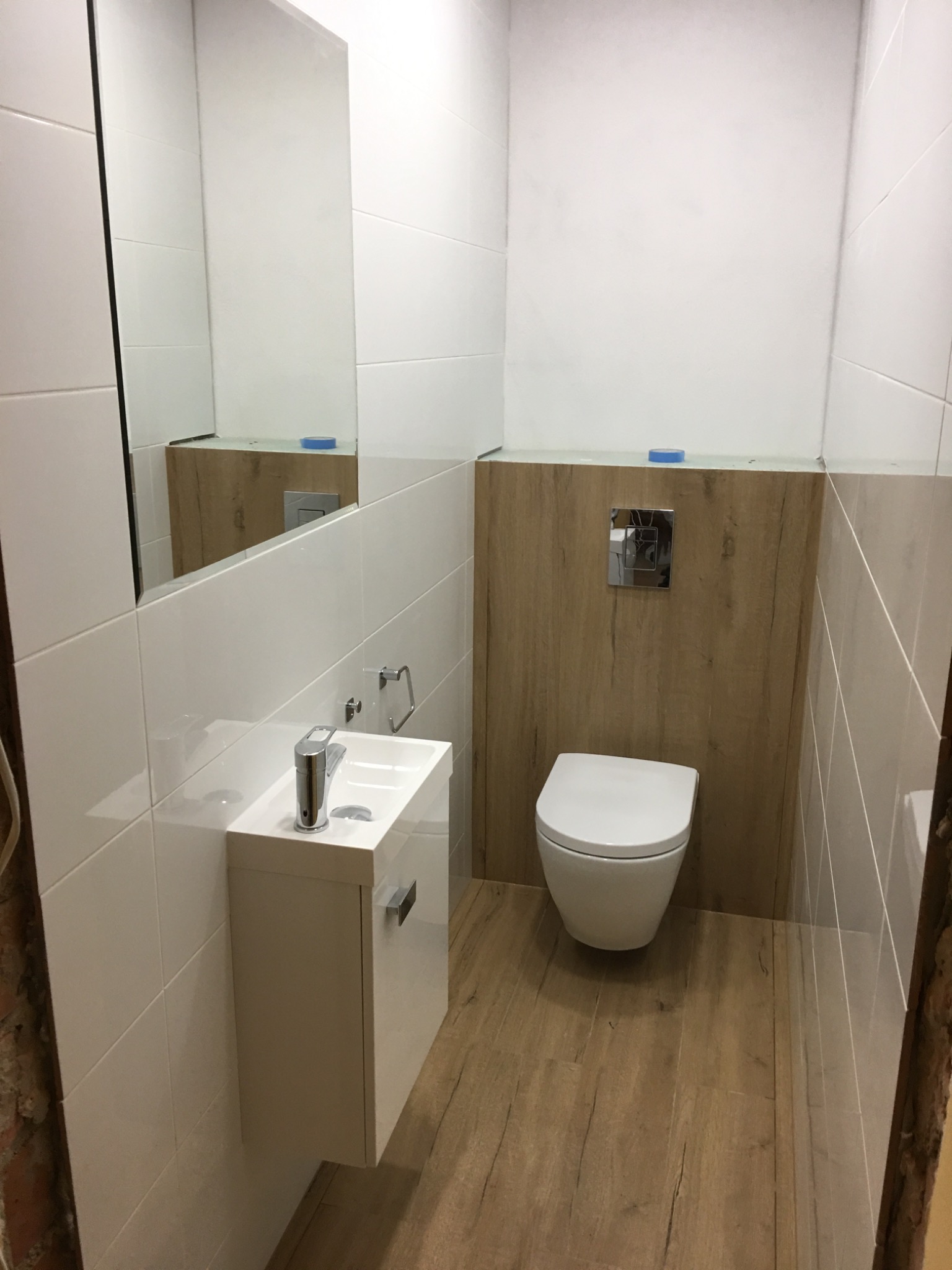 Wykończona łazienka w Bezrzeczu z wiszącą toaletą na tle drewnopodobnej okładziny, białą umywalką z szafką i lustrem, oraz podłogą imitującą drewno.