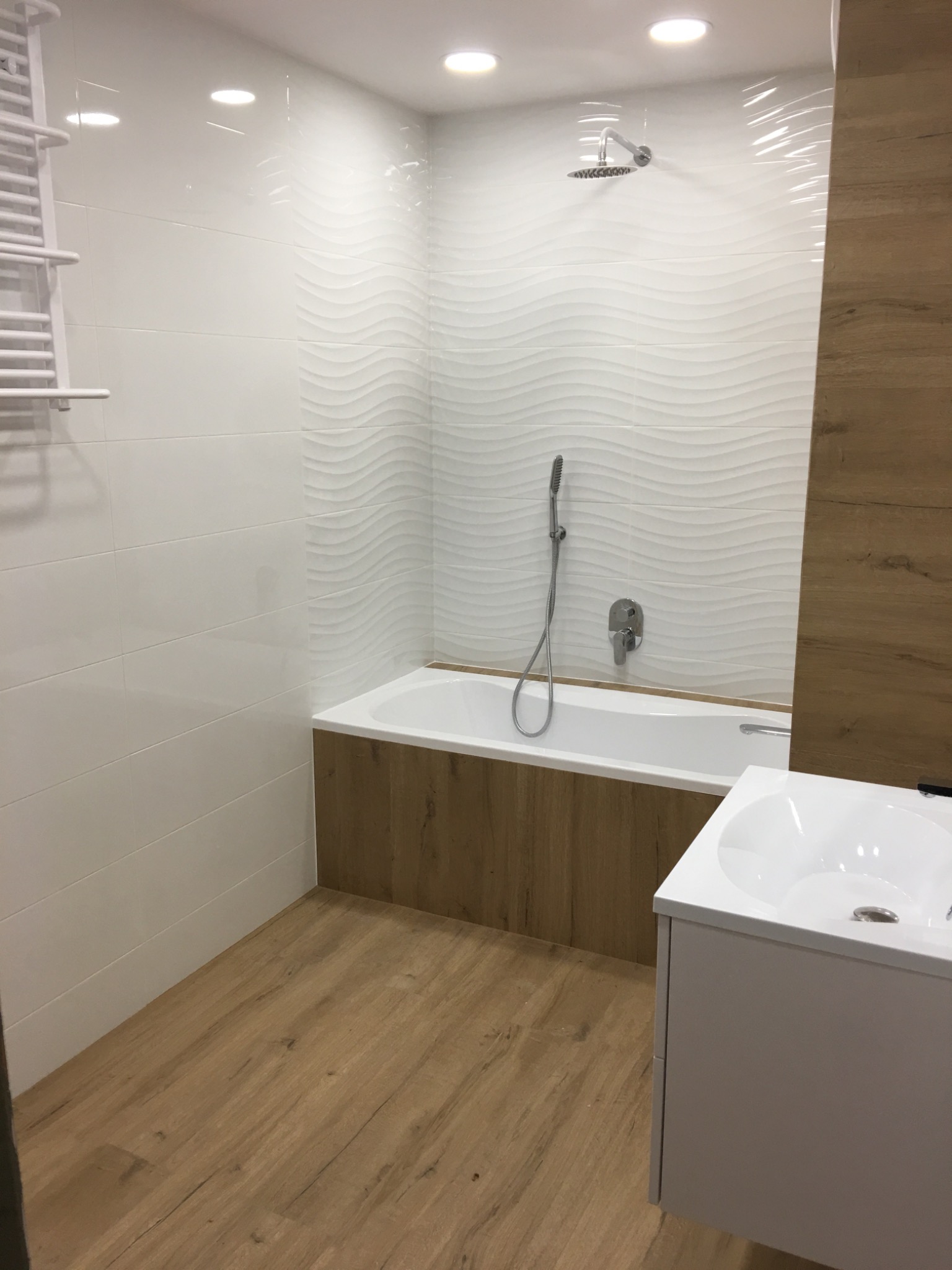 Nowoczesna łazienka po remoncie: białe płytki z falistym wzorem, drewnopodobne panele na podłodze i obudowie wanny, biała umywalka z szafką, chromowana bateria i deszczownica.