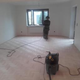 DARINSTAL Dariusz Zientara - Parkiet ułożony w jodełkę, szlifowany przez pracownika w słuchawkach, z widocznym odkurzaczem przemysłowym w pustym, jasnym pokoju.