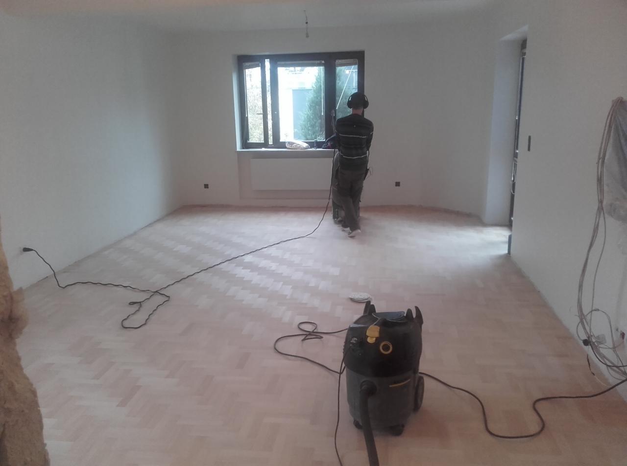 Parkiet ułożony w jodełkę, szlifowany przez pracownika w słuchawkach, z widocznym odkurzaczem przemysłowym w pustym, jasnym pokoju.