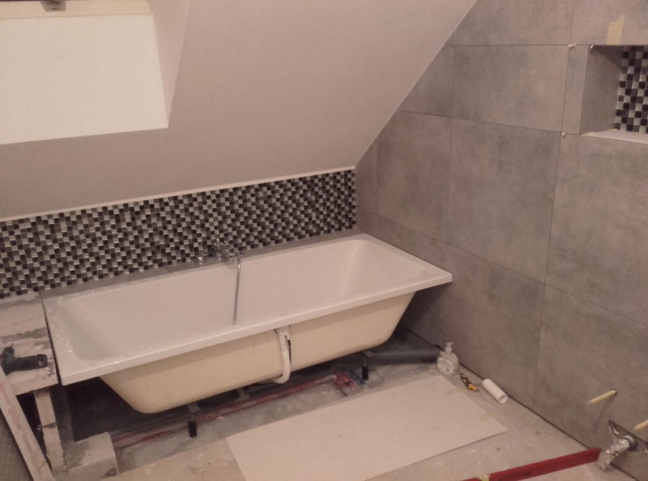 Wanna wolnostojąca z baterią, obłożona mozaiką i szarą płytką w trakcie montażu w poddaszowej łazience.