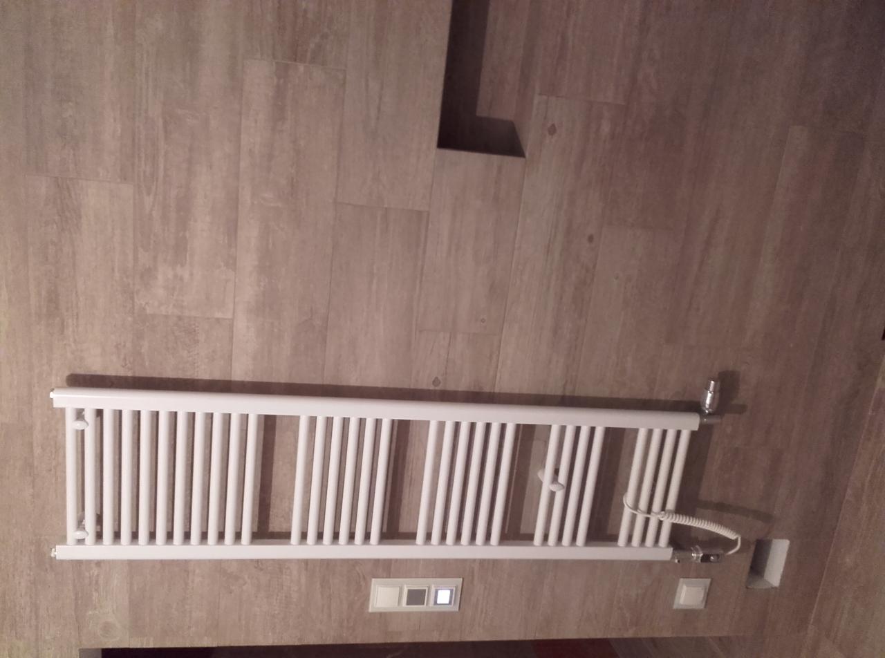 Biały grzejnik drabinkowy zamontowany poziomo na ścianie z płytek imitujących drewno, widoczne przyłącza i przewód zasilający.