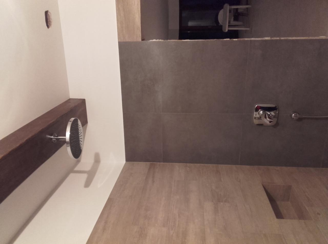 Nowoczesna łazienka z szarymi płytkami, drewnopodobną podłogą, deszczownicą zamontowaną na drewnianej belce i chromowaną baterią.