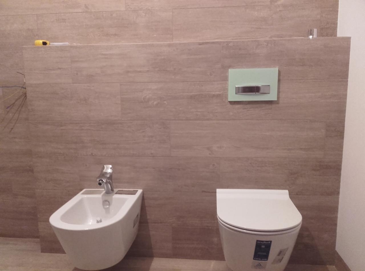 Nowoczesna łazienka z beżowymi płytkami imitującymi drewno, wiszący bidet i toaleta w kolorze białym, oraz zielony przycisk spłuczki.