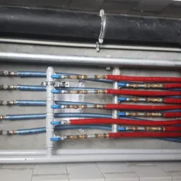 Rozdzielacz instalacji grzewczej z rurami PEX w kolorze czerwonym i niebieskim, z zaworami kulowymi i mosiężnymi złączkami, zamontowany na szarej ścianie w pomieszczeniu technicznym.