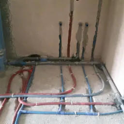 Surowe wnętrze z widocznymi rurami instalacji wodnej i grzewczej, częściowo wmurowanymi w ściany i ułożonymi na podłodze, z widocznymi mocowaniami i połączeniami.