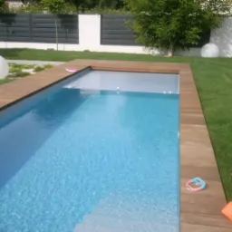 Prostokątny basen z drewnianym obramowaniem i częściowo zasłoniętą roletą, otoczony trawnikiem, z białymi kulami dekoracyjnymi i grillem w tle.