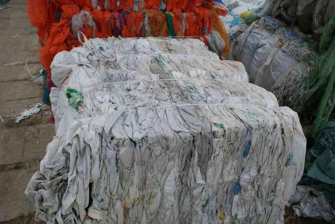 Stos sprasowanych, zużytych worków typu BIG BAG, związanych sznurkiem, z widocznymi zabrudzeniami i resztkami zawartości, obok sterty kolorowych siatek.