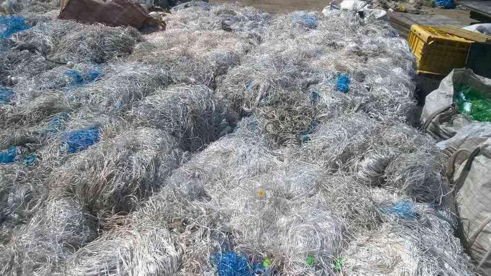 Stos poskręcanych, srebrnych drutów ze śladami niebieskiego plastiku, tworzących chaotyczny wzór na ziemi, z widocznymi elementami żółtej skrzynki i worka z zielonymi butelkami w tle.