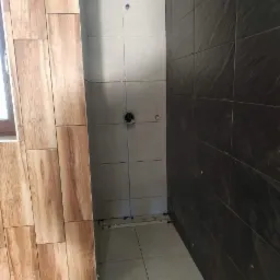 Wnętrze łazienki w trakcie prac wykończeniowych: ułożone płytki imitujące drewno na ścianie, jasne i czarne płytki na pozostałych ścianach, widoczne otwory na armaturę.