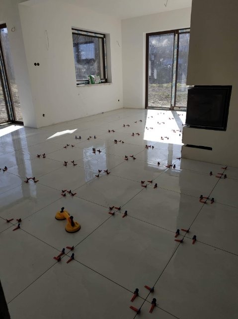 Układanie białych płytek podłogowych w jasnym pomieszczeniu, widoczne przyssawki do przenoszenia płytek i krzyżyki dystansowe, okna z widokiem na zewnątrz.