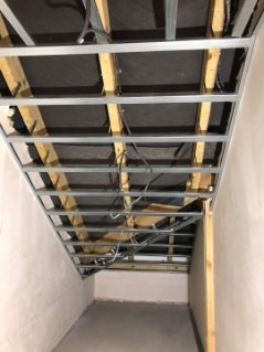 Montaż stelaża sufitu podwieszanego z widocznymi elementami konstrukcyjnymi drewnianymi i metalowymi, przewody elektryczne zwisają luźno, surowy stan wykończenia ścian i podłogi.