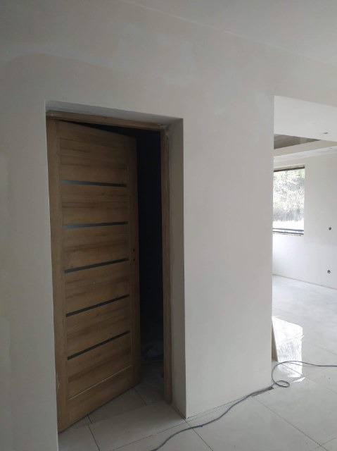 Świeżo wyszpachlowane ściany w jasnym pokoju z drewnianymi drzwiami z ciemnymi poziomymi paskami, widoczne ślady szpachlowania na ścianach i podłodze.