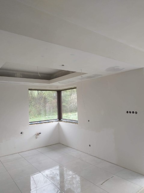 Pokój w trakcie remontu: białe ściany po szpachlowaniu, widoczne niedoskonałości i otwory na gniazdka elektryczne, biała podłoga z płytek, okno narożne z widokiem na zieleń, sufit z obniżonym...