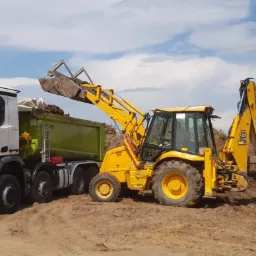 Żółta koparko-ładowarka JCB nabiera ziemię do zielonej ciężarówki Mercedes-Benz na placu budowy w słoneczny dzień.
