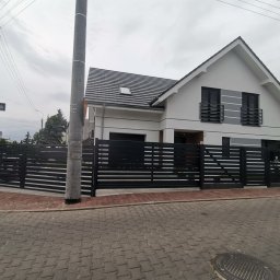 Cr-budsystem Robert Cendalski - Nowoczesny, czarny płot panelowy z poziomych profili, otaczający posesję z białym domem z szarą dachówką, widok z ulicy wyłożonej kostką.