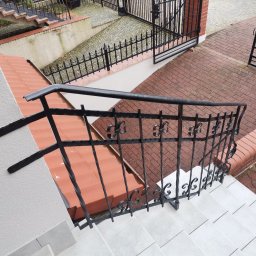 Cr-budsystem Robert Cendalski - Kute balustrady schodowe z dekoracyjnymi elementami, widok z góry na białe płytki schodowe i czerwoną cegłę na posesji, brama wjazdowa z kutego żelaza w tle.
