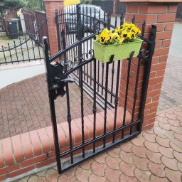 Cr-budsystem Robert Cendalski - Otwarta, czarna furtka z metalu, umieszczona między dwoma ceglanymi słupkami, z zielonym pojemnikiem z żółtymi bratkami, na tle brukowanej ścieżki i zieleni.