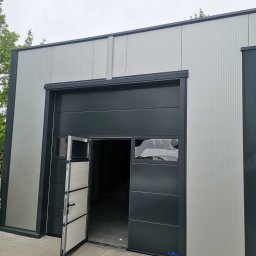 Cr-budsystem Robert Cendalski - Otwarta segmentowa brama przemysłowa w kolorze grafitowym z oknami, zamontowana w budynku o srebrnej, ryflowanej elewacji.