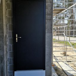 Cr-budsystem Robert Cendalski - Czarna, metalowa, zewnętrzna drzwi z srebrną klamką, zamontowana w surowej, betonowej ścianie budynku w trakcie budowy, widoczne rusztowanie i teren budowy w tle.