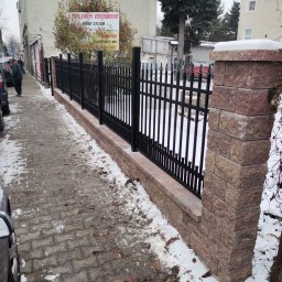 Cr-budsystem Robert Cendalski - Czarne, metalowe ogrodzenie z ceglanymi słupkami i podmurówką, częściowo pokryte śniegiem, wzdłuż chodnika w miejskiej scenerii.