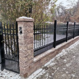 Cr-budsystem Robert Cendalski - Nowoczesne ogrodzenie z czarnych paneli metalowych i słupków z cegły klinkierowej, częściowo pokryte śniegiem, widoczne w zimowej scenerii.