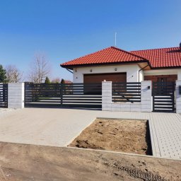 Cr-budsystem Robert Cendalski - Nowoczesna, pozioma, czarna brama wjazdowa z przesuwnym skrzydłem i furtką, osadzona na murowanych, szarych słupkach z betonu architektonicznego, na tle domu z czerwoną dachówką i podjazdem...