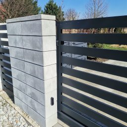 Cr-budsystem Robert Cendalski - Nowoczesna, pozioma brama w kolorze antracytowym z betonowym słupkiem z bloczków, widoczny czytnik zbliżeniowy, kostka brukowa i biały żwir u podstawy.