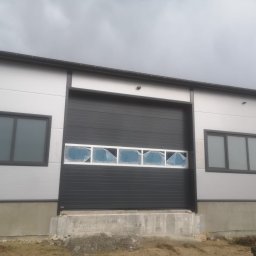 Cr-budsystem Robert Cendalski - Szara brama segmentowa z oknami zabezpieczonymi folią ochronną, zamontowana w hali przemysłowej z szarą elewacją i oknami, widok z zewnątrz w pochmurny dzień.