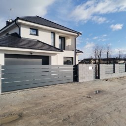 Cr-budsystem Robert Cendalski - Nowoczesny dom z szarą bramą przesuwną i ogrodzeniem z betonowych bloczków, widoczne kałuże na gruncie.