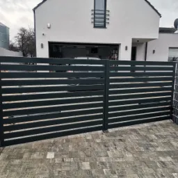 Nowoczesna, pozioma, czarna brama wjazdowa na posesję z białym domem w tle, widoczny silos i ogrodzenie gabionowe po prawej stronie.