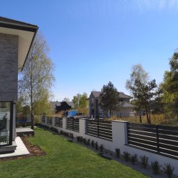 Cr-budsystem Robert Cendalski - Nowoczesne ogrodzenie panelowe z poziomymi przęsłami w kolorze antracytowym, osadzone na białych, murowanych słupkach, otoczone świeżo posadzoną roślinnością na tle zielonego trawnika i domów...