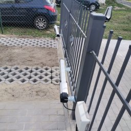 Cr-budsystem Robert Cendalski - Szara, metalowa brama przesuwna z automatyką, widoczne elementy montażowe i silnik. W tle kostka ażurowa i dwa samochody osobowe