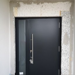 Cr-budsystem Robert Cendalski - Ciemne drzwi zewnętrzne z szybą boczną i długim uchwytem, zamontowane w surowym otworze z widocznymi warstwami izolacji i tynku.