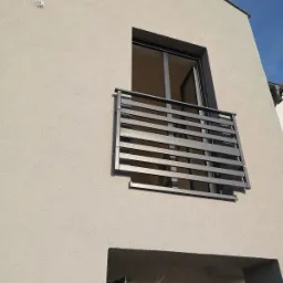 Nowoczesny balkon z szarymi, poziomymi barierkami na tle jasnej elewacji budynku i błękitnego nieba.