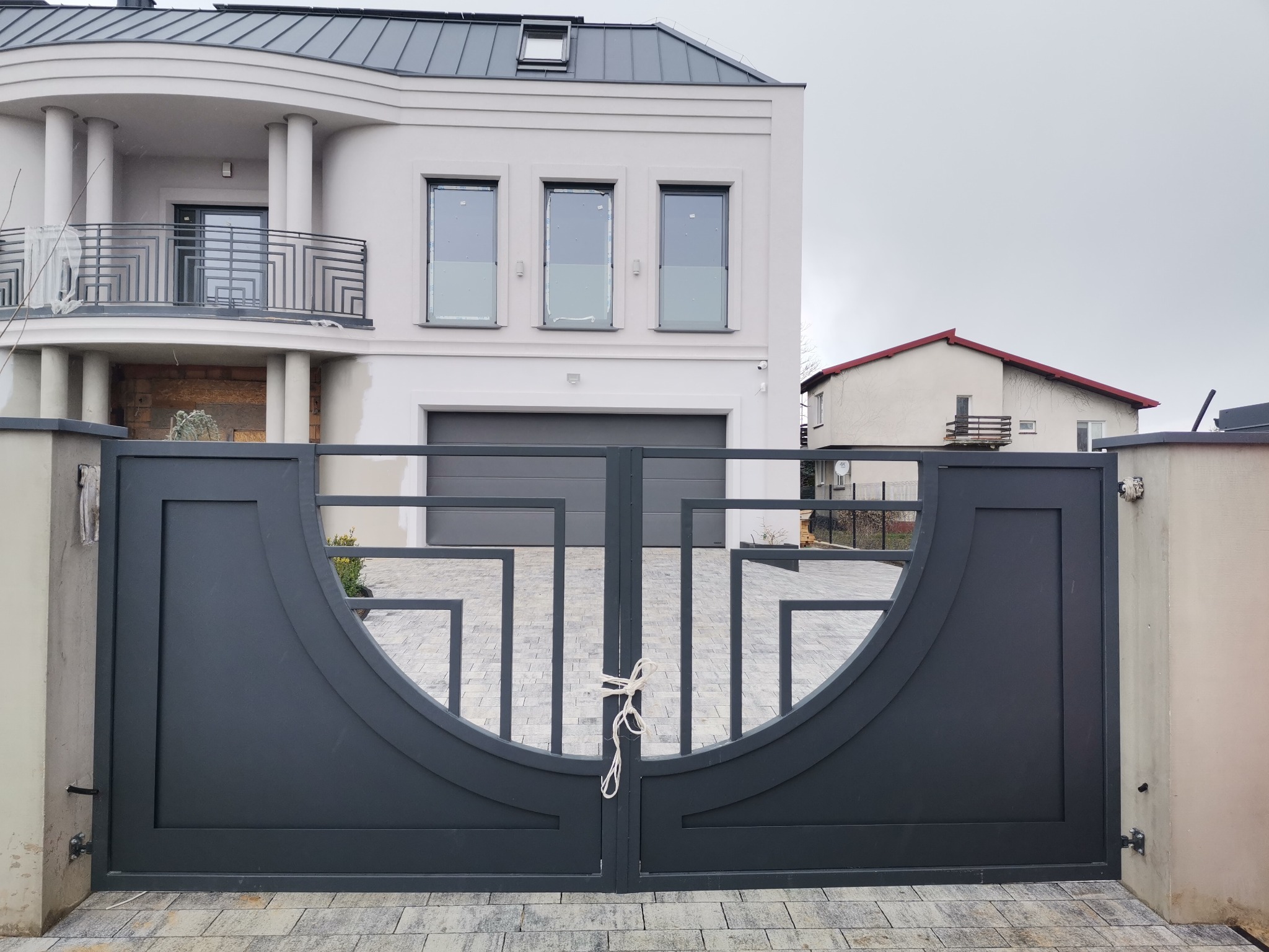 Nowoczesna, grafitowa brama wjazdowa z geometrycznym wzorem, tymczasowo zabezpieczona sznurkiem, na tle domu z szarą stolarką okienną i garażową.