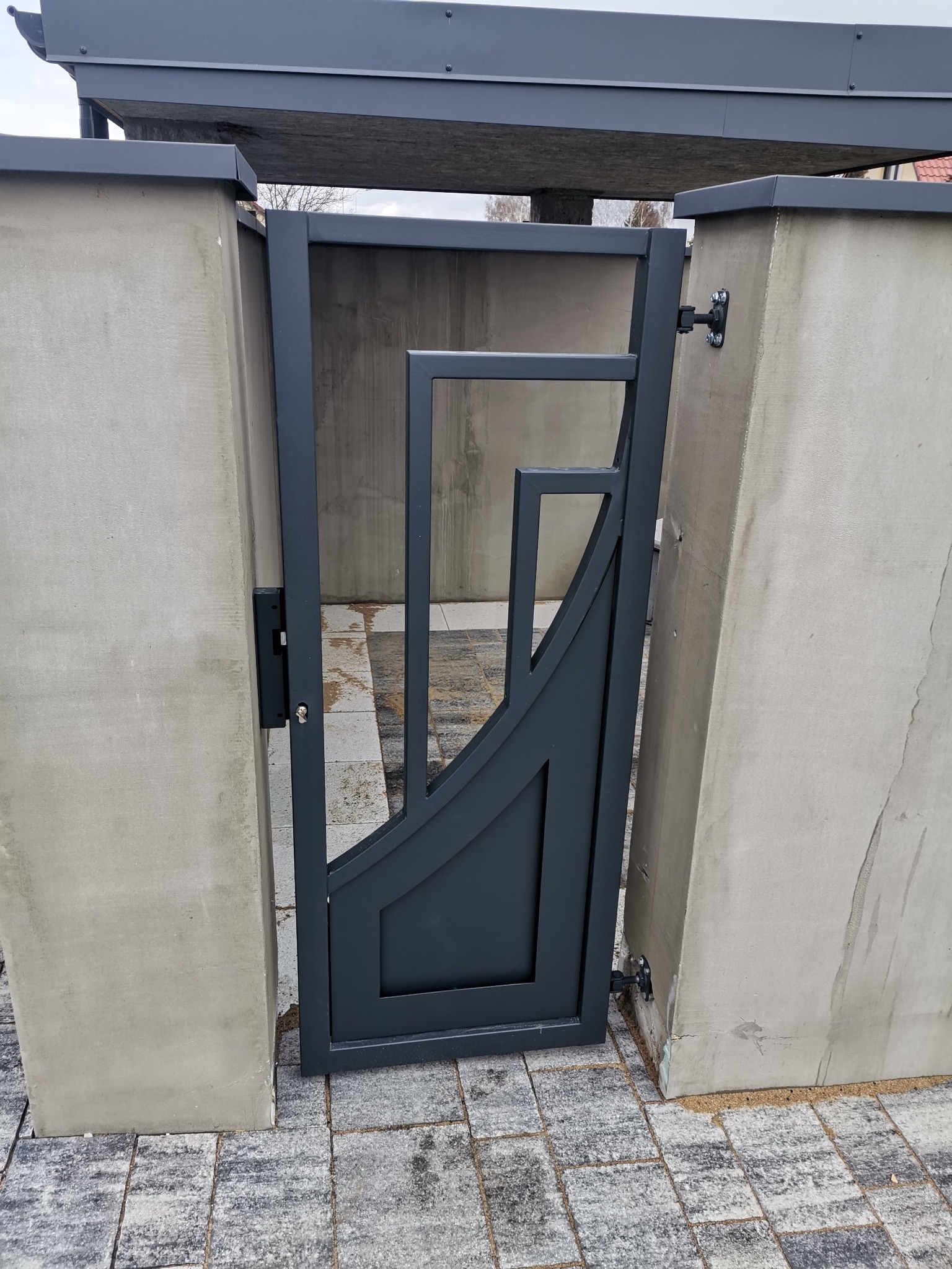 Nowoczesna furtka ogrodzeniowa z geometrycznym wzorem, zamontowana między betonowymi słupkami na kostce brukowej.
