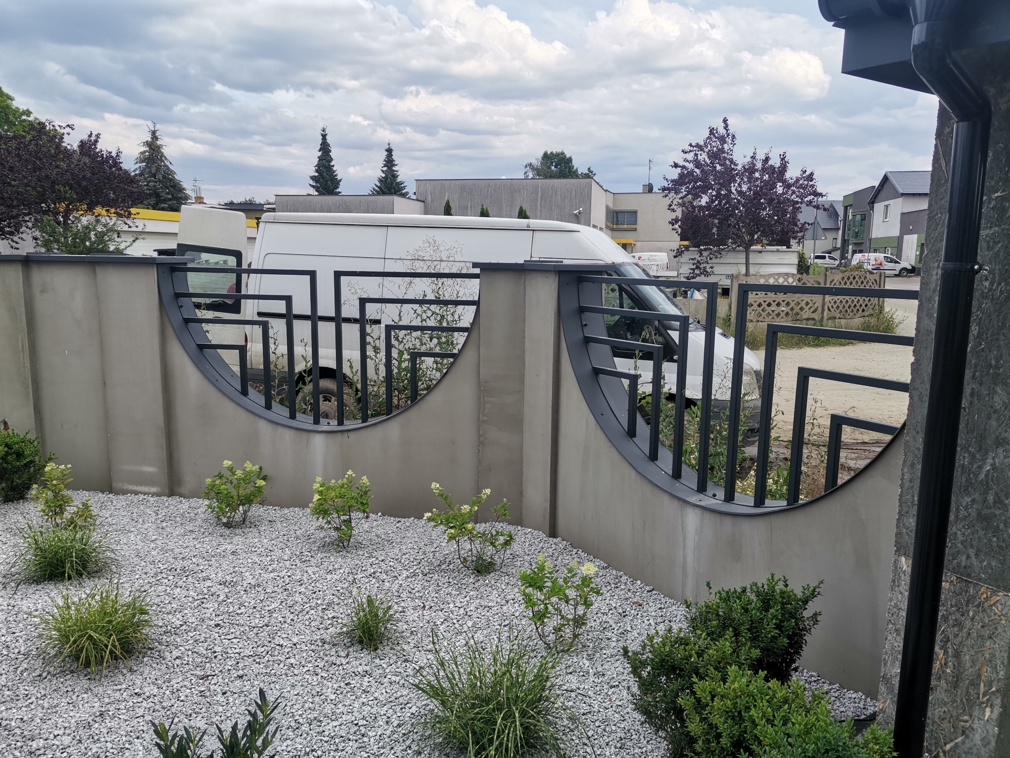 Nowoczesne ogrodzenie betonowe z metalowymi przęsłami o półokrągłym kształcie, widok na biały samochód dostawczy za ogrodzeniem, szara nawierzchnia z kamyczków i roślinność.