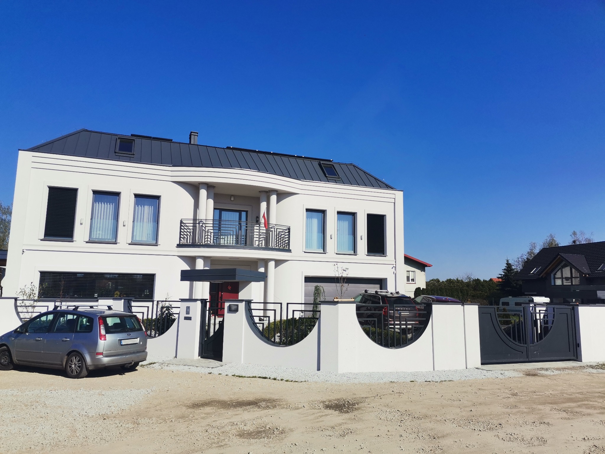 Nowoczesny dom jednorodzinny z białymi ścianami i szarą dachówką, otoczony niskim, białym ogrodzeniem z metalowymi przęsłami i furtką, widok z zewnątrz, samochód zaparkowany przed domem.