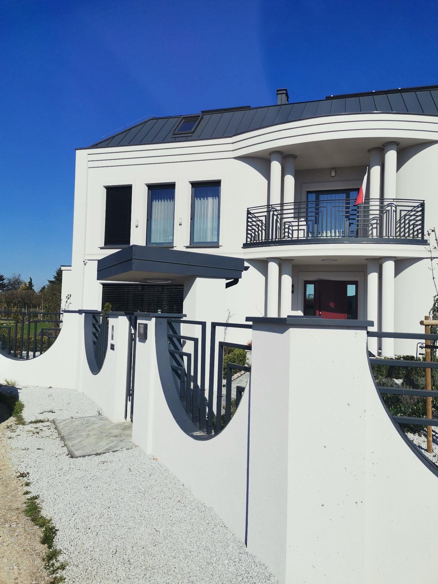 Nowoczesny dom jednorodzinny z białą elewacją, szarym dachem i minimalistycznym ogrodzeniem z elementami metalowymi. Widoczny podjazd wysypany białym żwirem i balkon z metalową balustradą.
