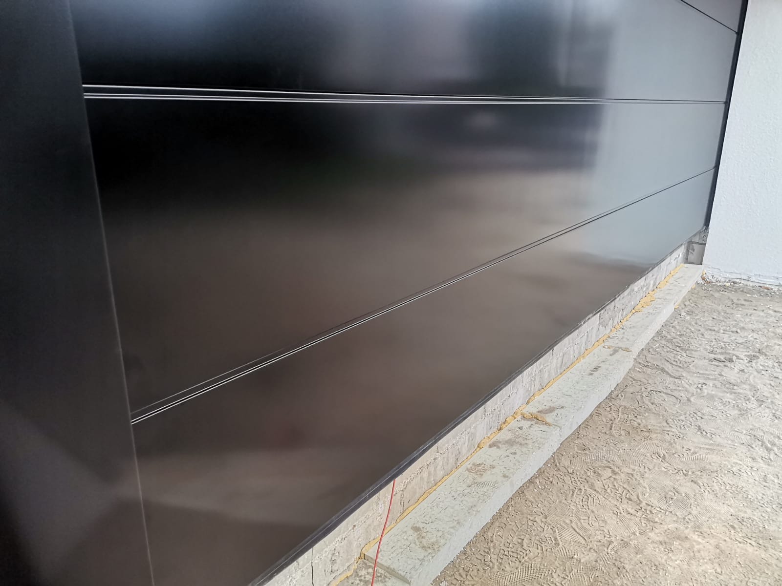 Fragment czarnej, segmentowej konstrukcji z poziomymi, srebrnymi liniami, prawdopodobnie w trakcie montażu, widoczne uszczelnienie silikonowe na betonowej wylewce.