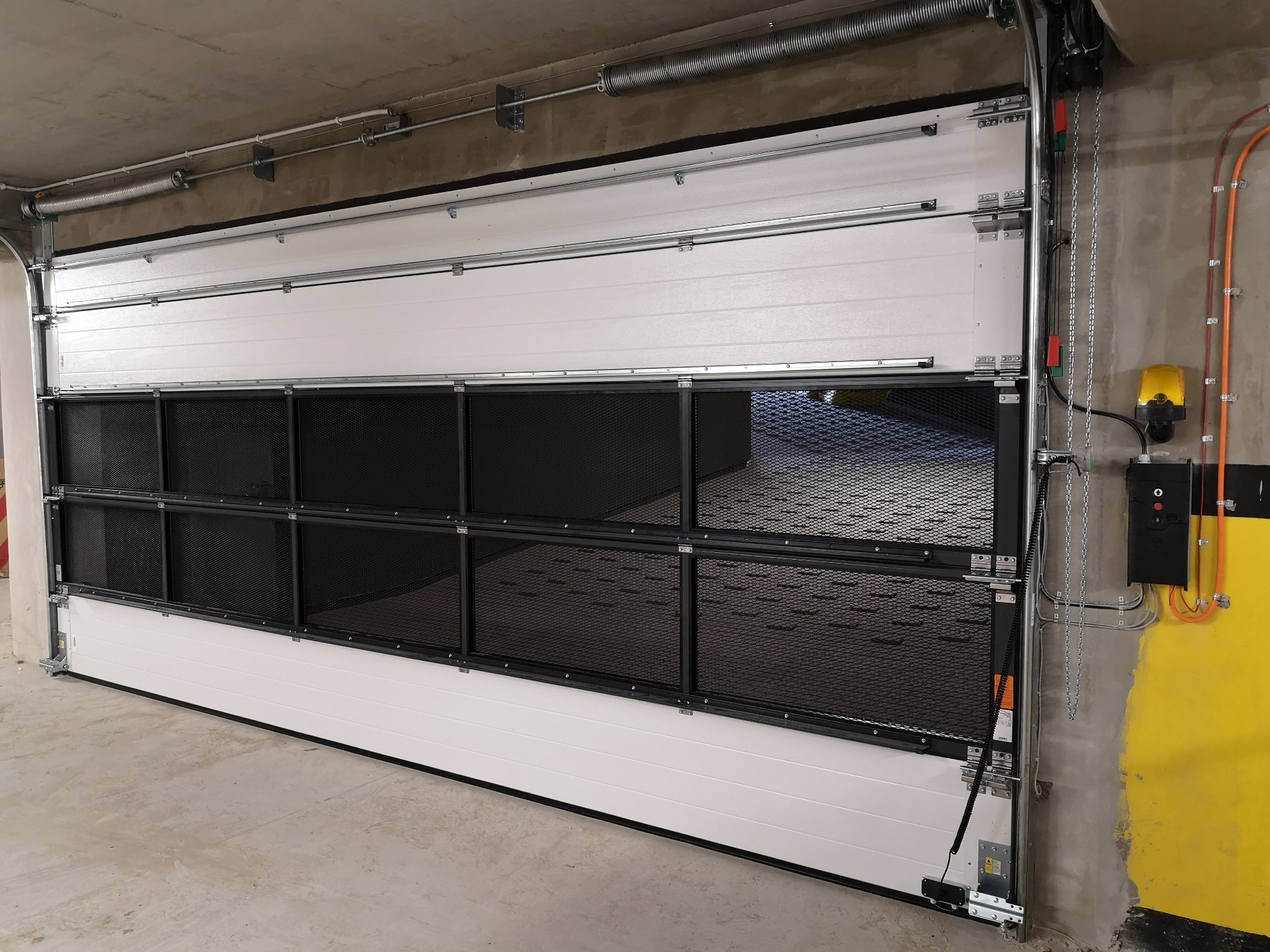 Zamknięta, segmentowa brama z kratką wentylacyjną w garażu podziemnym, widoczne sprężyny i mechanizm otwierania.