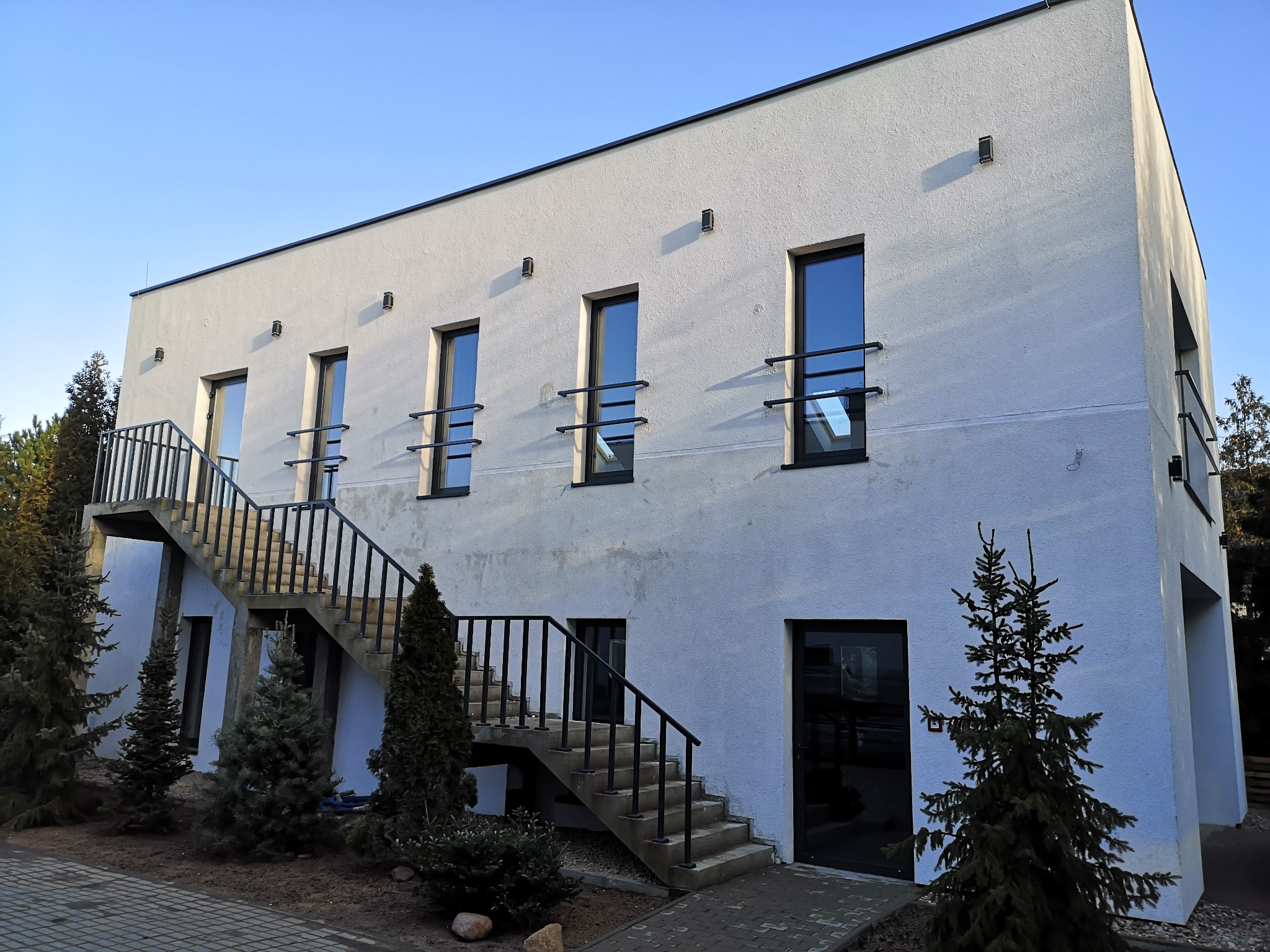 Nowoczesny budynek z białym tynkiem, czarnymi oknami i zewnętrznymi schodami z balustradą w kolorze antracyt, otoczony zielenią iglaków.