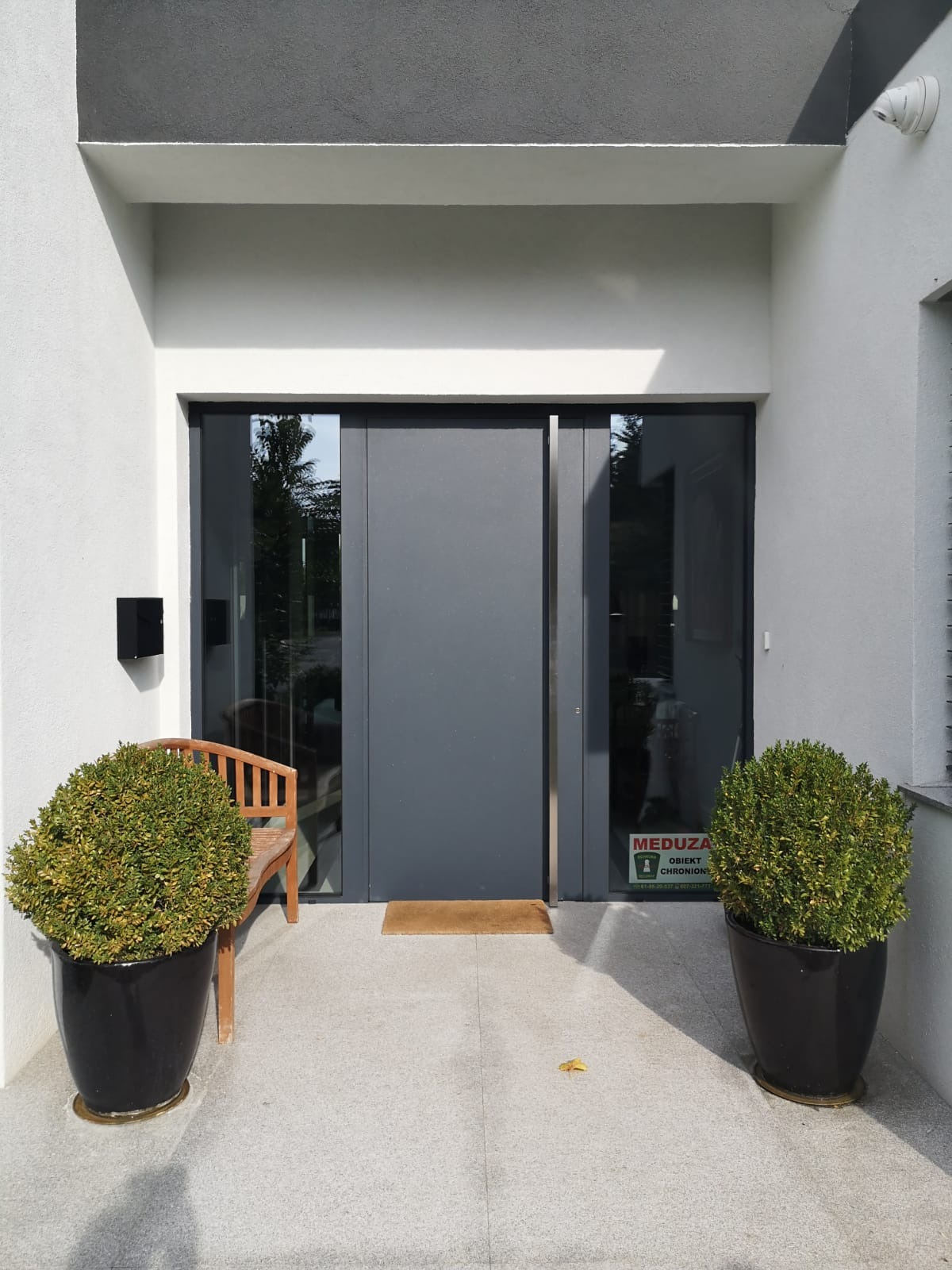 Nowoczesne szare drzwi zewnętrzne z przeszkleniem bocznym i naklejką 'Obiekt Chroniony', usytuowane we wnęce budynku, z donicami z bukszpanem po bokach i małą ławką z lewej strony.