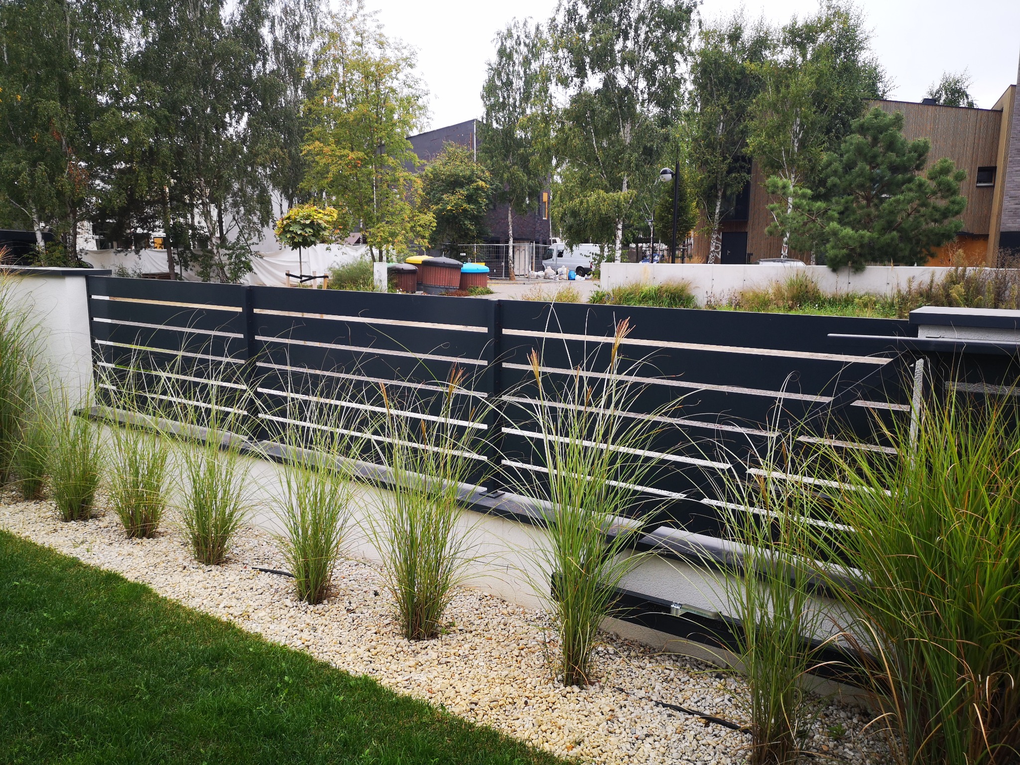 Nowoczesne ogrodzenie panelowe z poziomymi przerwami, obsadzone ozdobnymi trawami na białym żwirze, w tle drzewa i budynek.