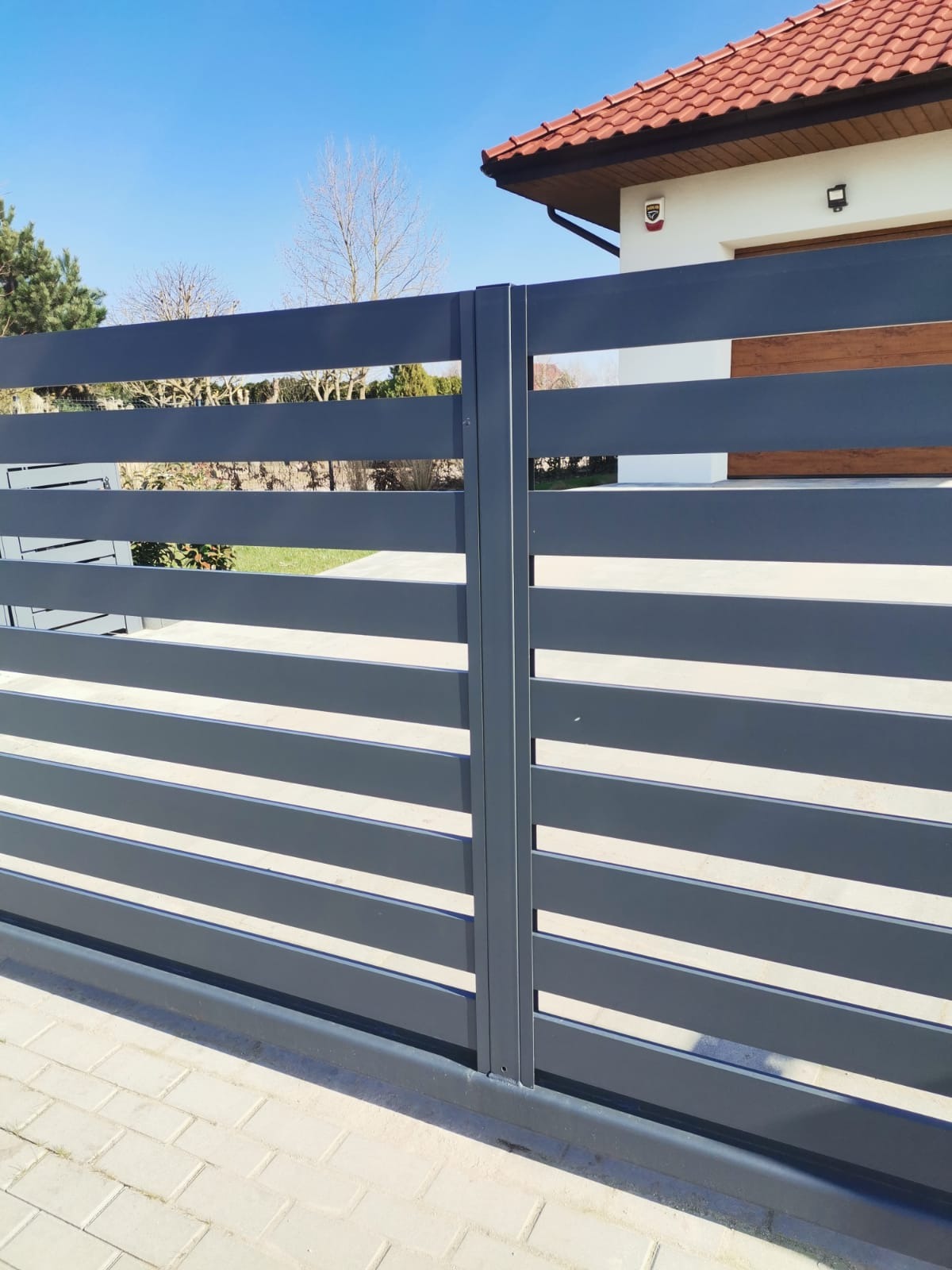 Nowoczesna brama wjazdowa z poziomych, szarych profili, widok z zewnątrz na tle domu z czerwoną dachówką i jasną elewacją.